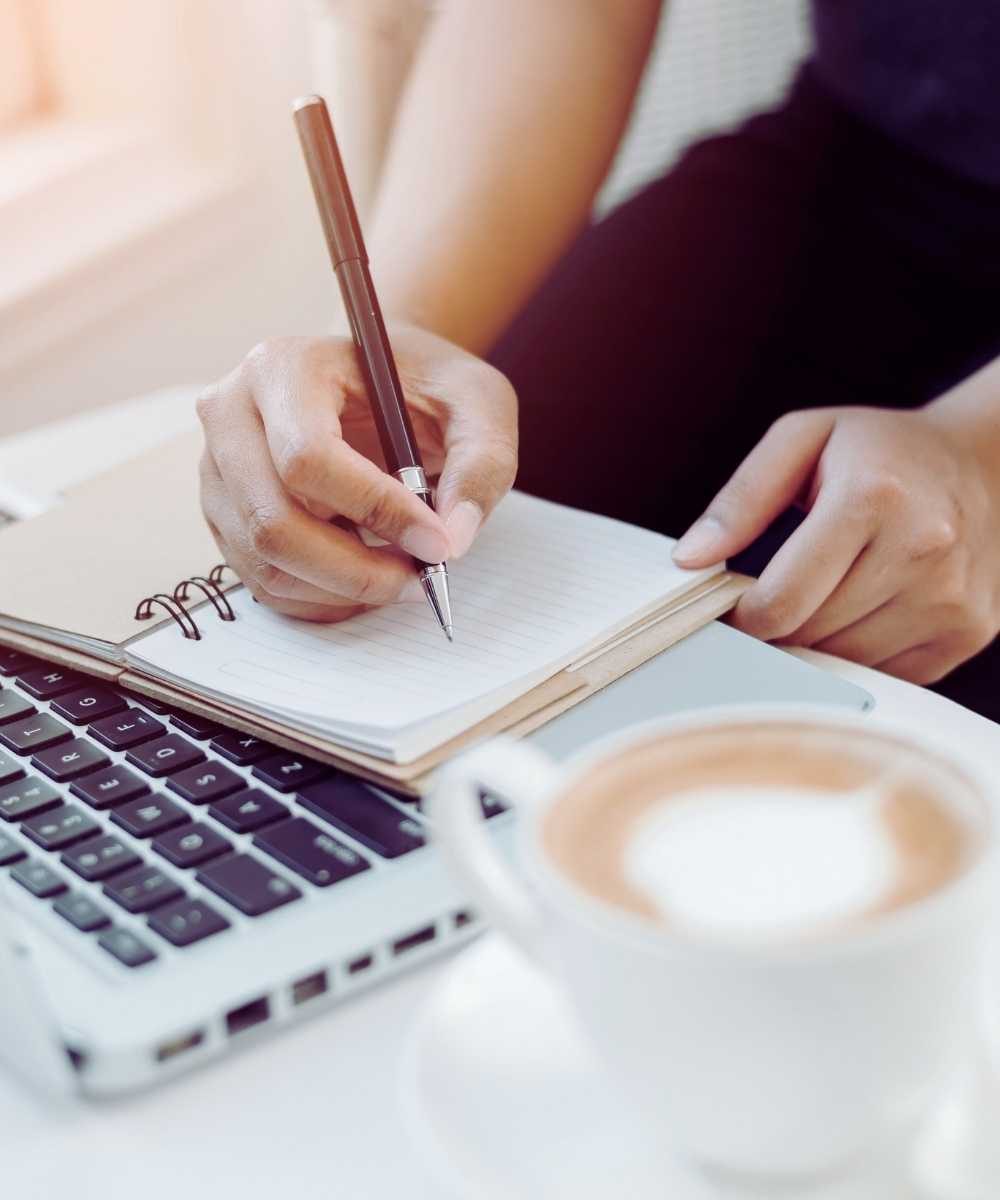Woman working on laptop and writing in journal with coffee